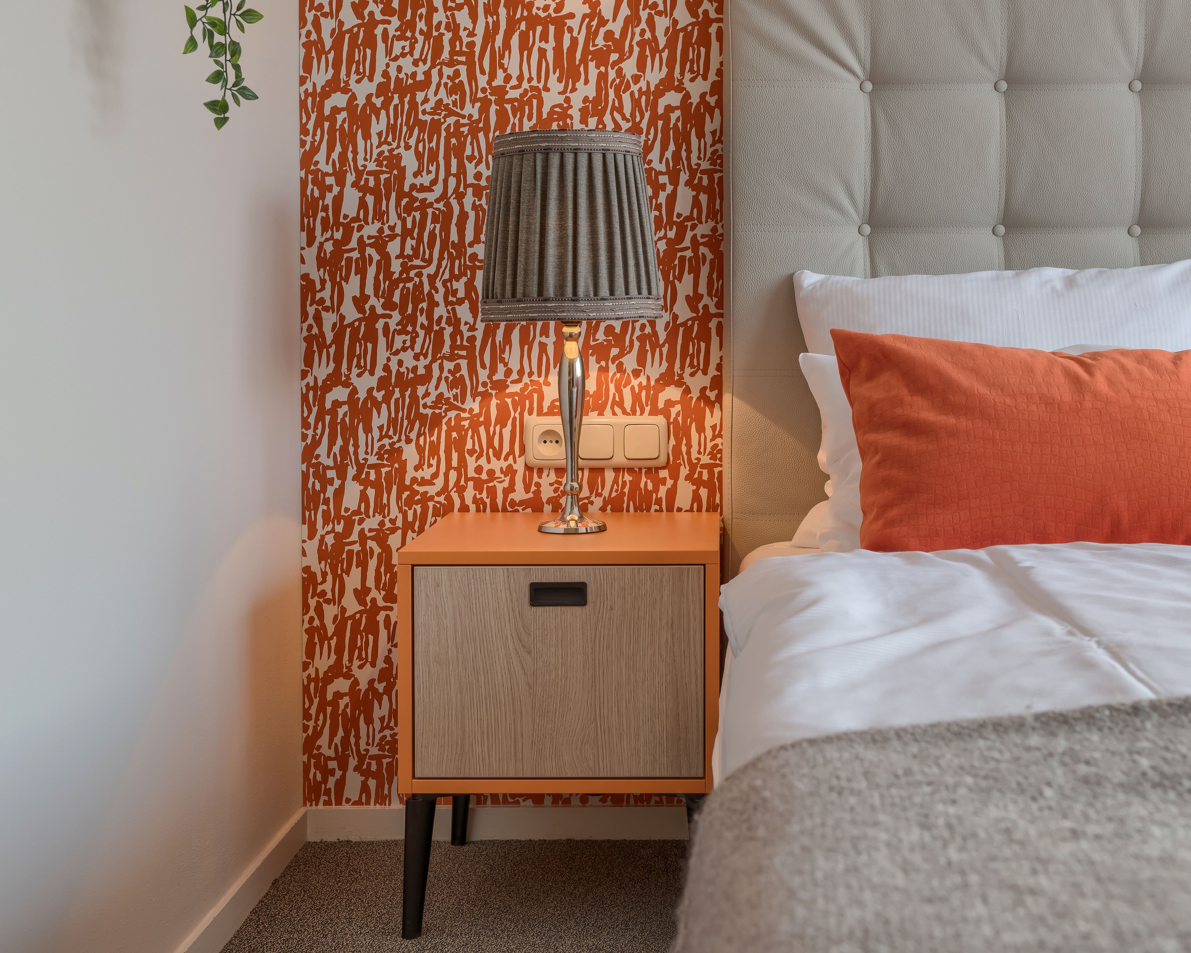 A neatly made bed with white and orange pillows, a grey headboard, and a bedside lamp on a wooden nightstand.