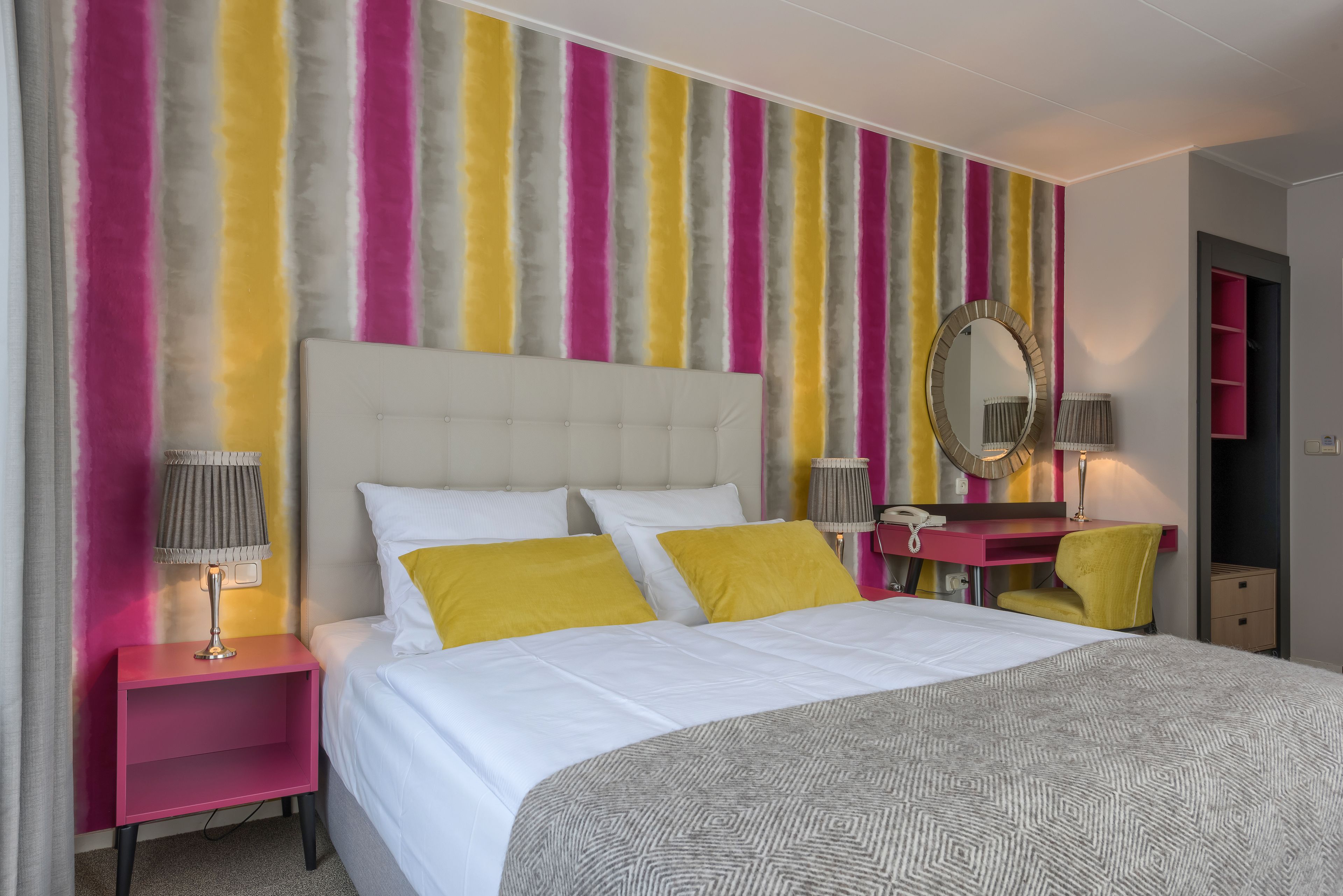 Una camera d'albergo con un letto grande, un comodino con una lampada, una toeletta con uno specchio e una parete colorata a righe.