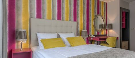 A hotel room with a large bed, a bedside table with a lamp, a vanity with a mirror, and a colorful striped wall.