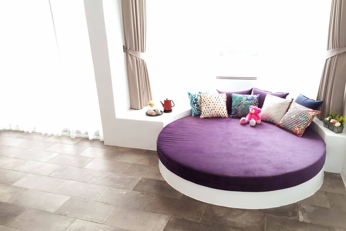 Una cama redonda con un colchón morado en una habitación luminosa con un ventanal grande, paredes blancas y suelo de baldosas.