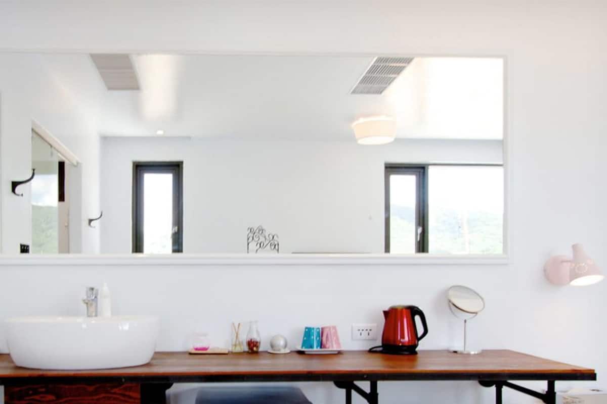 Un baño moderno con un espejo grande, un lavabo blanco, un mueble de tocador de madera y una tetera roja.