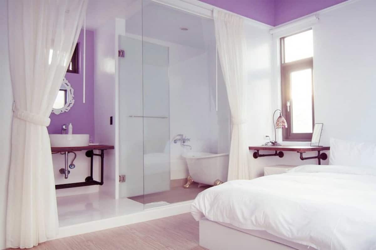 Un dormitorio con cama, mesita de noche, espejo, ventana y baño con lavabo y ducha.