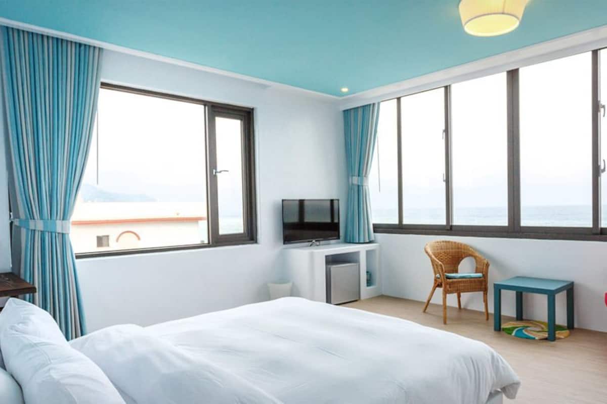 Habitación de hotel con cama, televisión, silla, una mesita y ventanas amplias con cortinas.