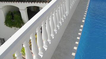 Zona junto a la piscina con barandilla blanca, piscina azul y vista a un edificio con tejado de azulejos y una guirnalda.