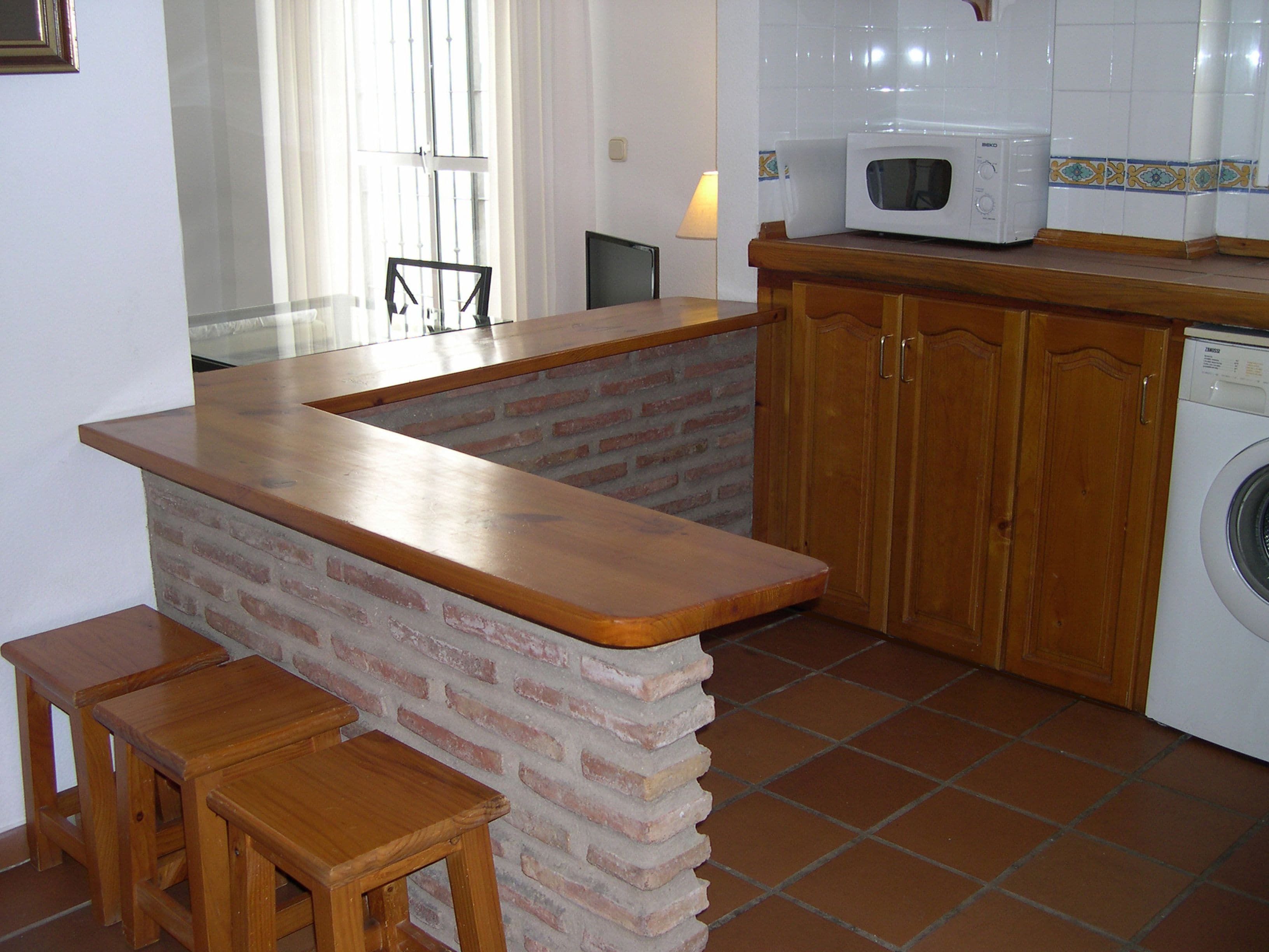 A compact kitchen with a wooden countertop, a brick accent wall, a washing machine, and wooden cabinets.