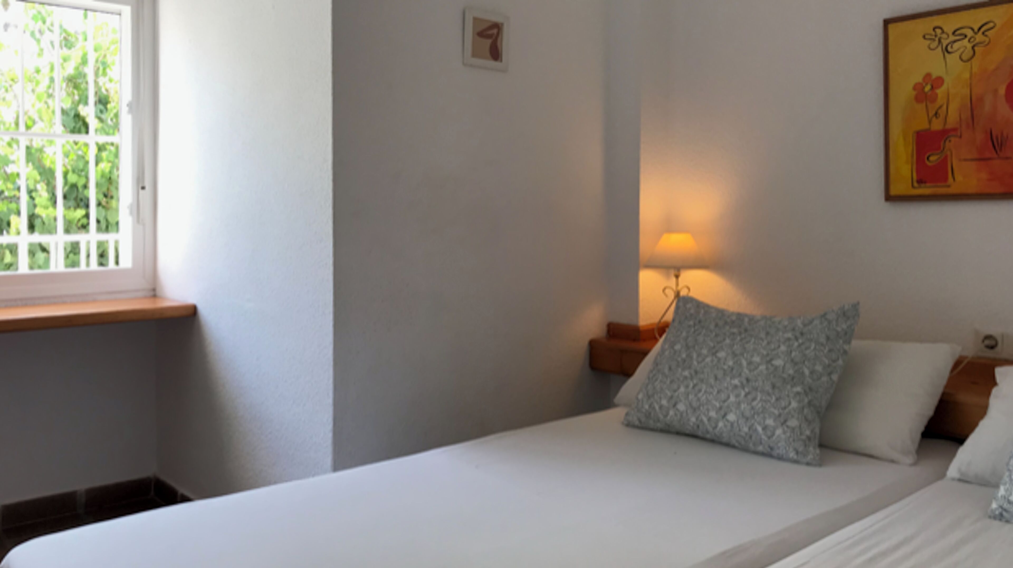 Ein Schlafzimmer mit einem Bett, einem Fenster, einem Nachttisch mit Lampe und einem Wandbild.