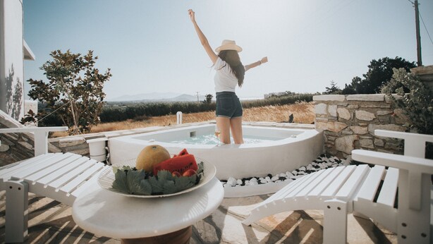 A person standing in a hot tub with arms raised, surrounded by white lounge chairs and a table with fruit.
