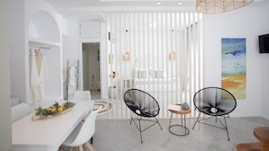 A modern, well-lit room with a dining area featuring a white table and two black chairs, a wicker pendant light, and a painting on the wall.