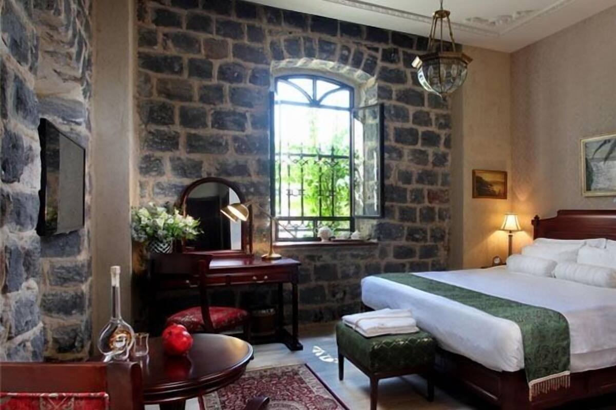 Une chambre avec un mur en pierre, une grande fenêtre, un lit avec du linge de maison blanc, un bureau avec une lampe et une chaise.