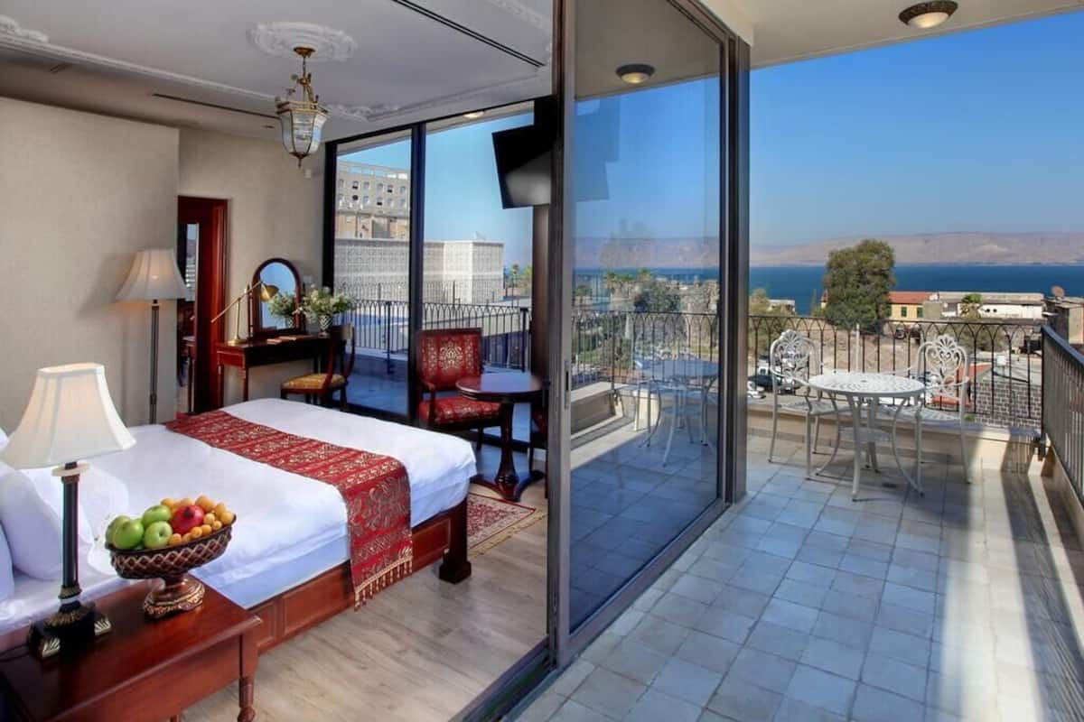 Une chambre d’hôtel avec un lit, une table de chevet et un panier de fruits. La chambre dispose d’un balcon avec vue sur la mer et les bâtiments.
