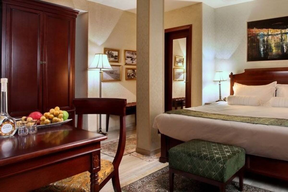 Une chambre d’hôtel avec un grand lit, un bureau en bois, une chaise, une petite table avec des fruits et une bouteille, et un tableau accroché au mur.