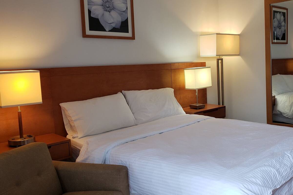 A neatly made bed with white linens, two bedside lamps, a wooden headboard, a chair, and a framed flower on the wall.