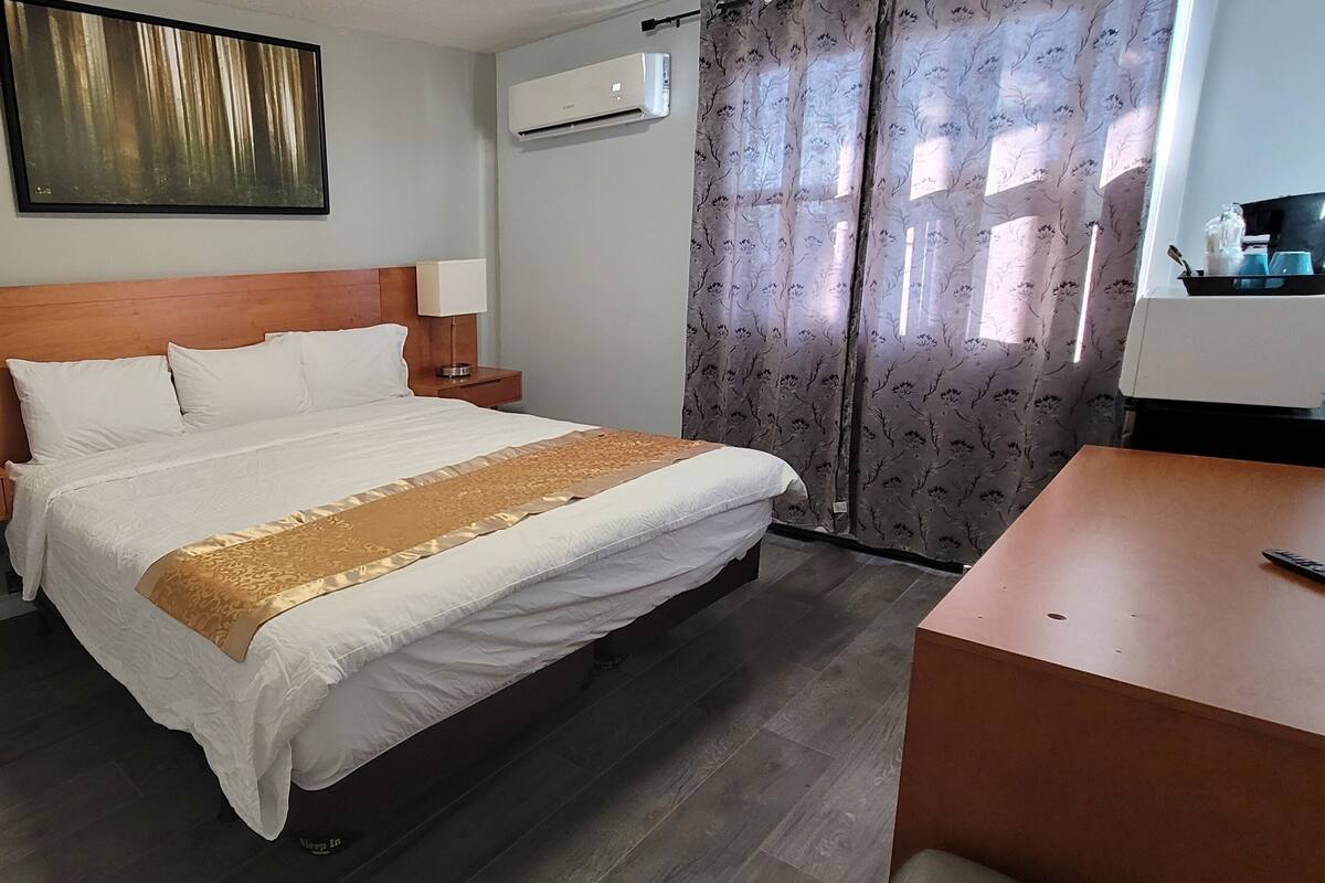 A hotel room with a wooden headboard, a bed with white linens and a gold accent, a nightstand with a lamp, an air conditioning unit, and a desk with a chair.