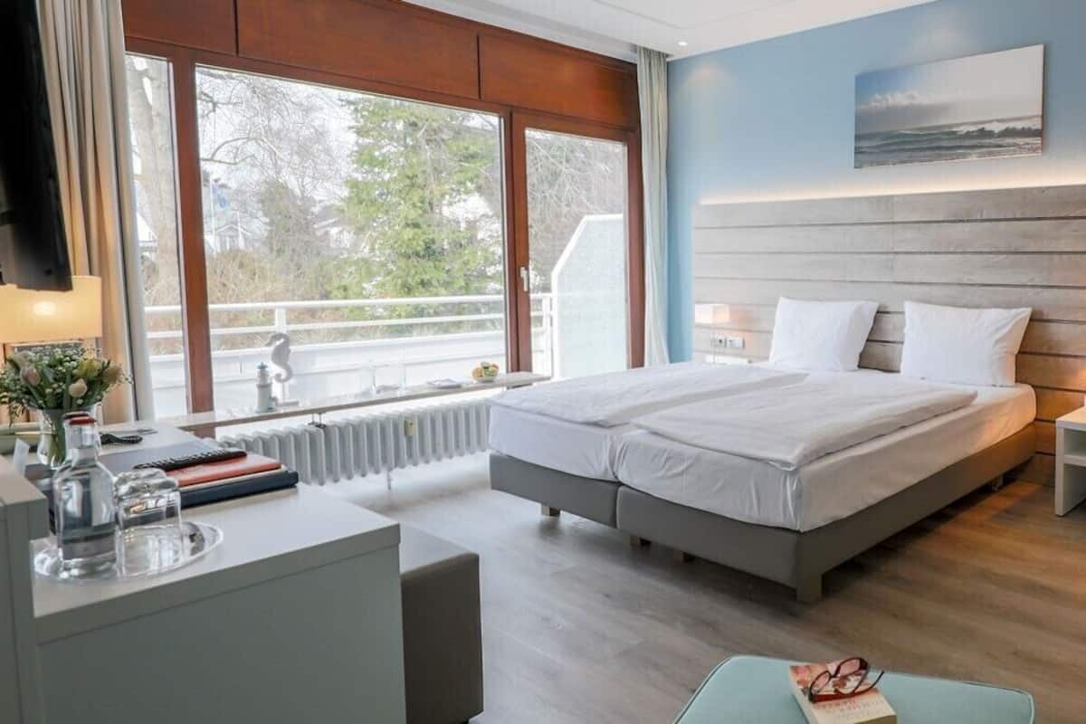 A modern hotel room with a large bed, bedside tables, a desk with a chair, and a view of the outdoors through large windows.