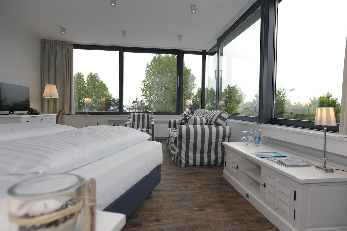 A modern hotel room with a large bed, a seating area with striped chairs, a desk with a lamp, and large windows offering a view of trees.