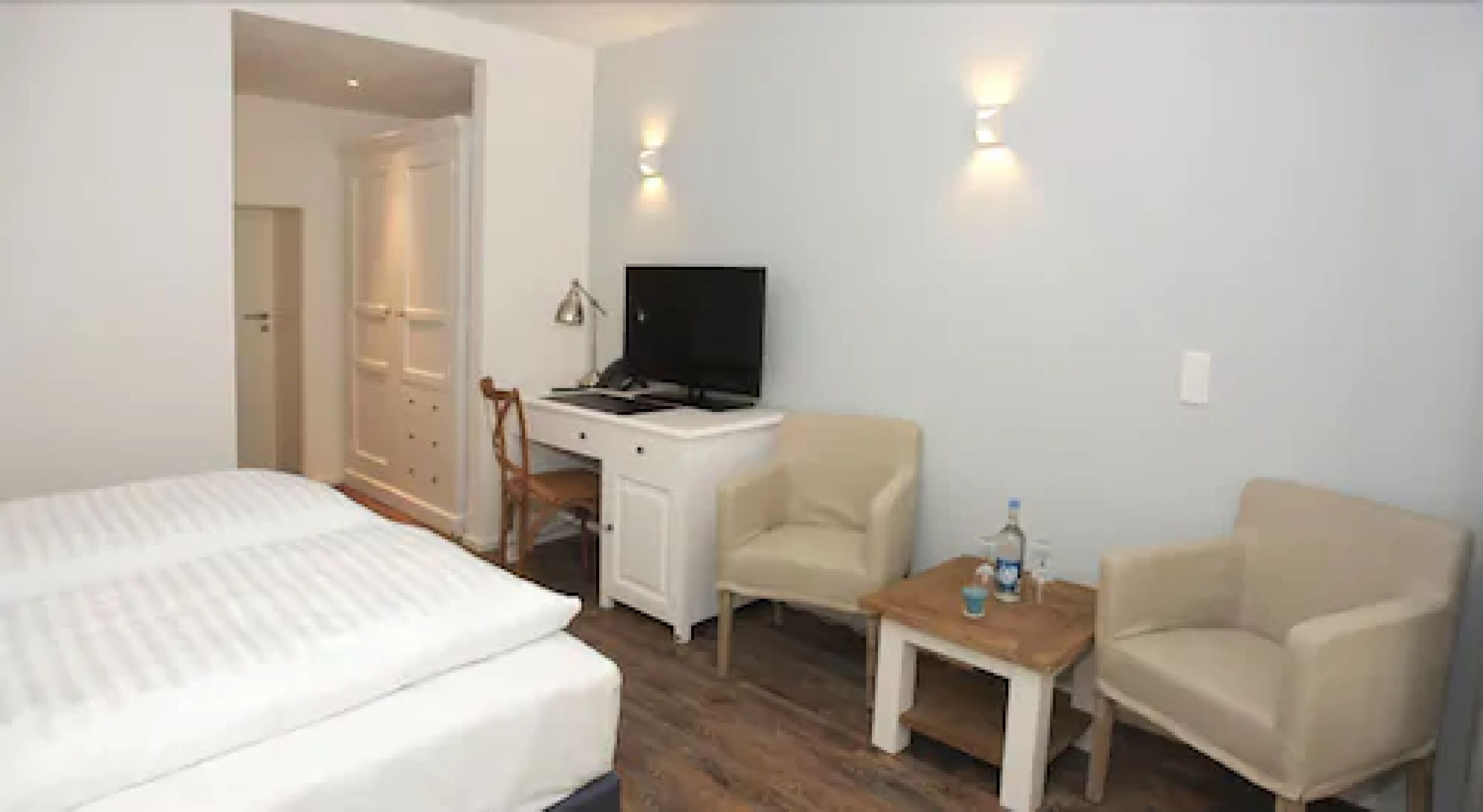 Ein Hotelzimmer mit einem Bett, einem Schreibtisch mit Computer, zwei Stühlen und einem kleinen Tisch mit einer Flasche.