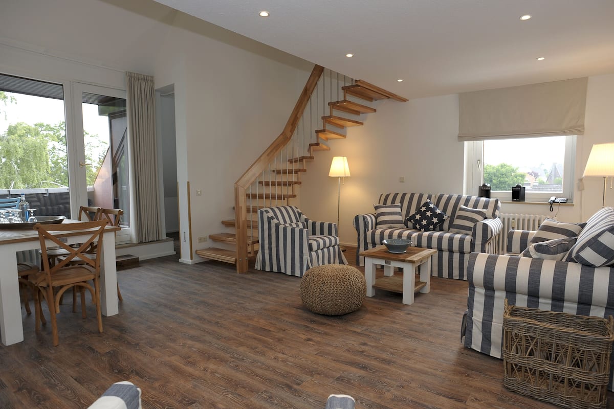 A spacious living room with a wooden staircase, striped sofas, and a dining area with wooden chairs.