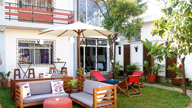 A patio area with a wooden sofa, red chairs, and a table. There are potted plants and a large umbrella providing shade. - Villa Mahefa (Antananarivo)