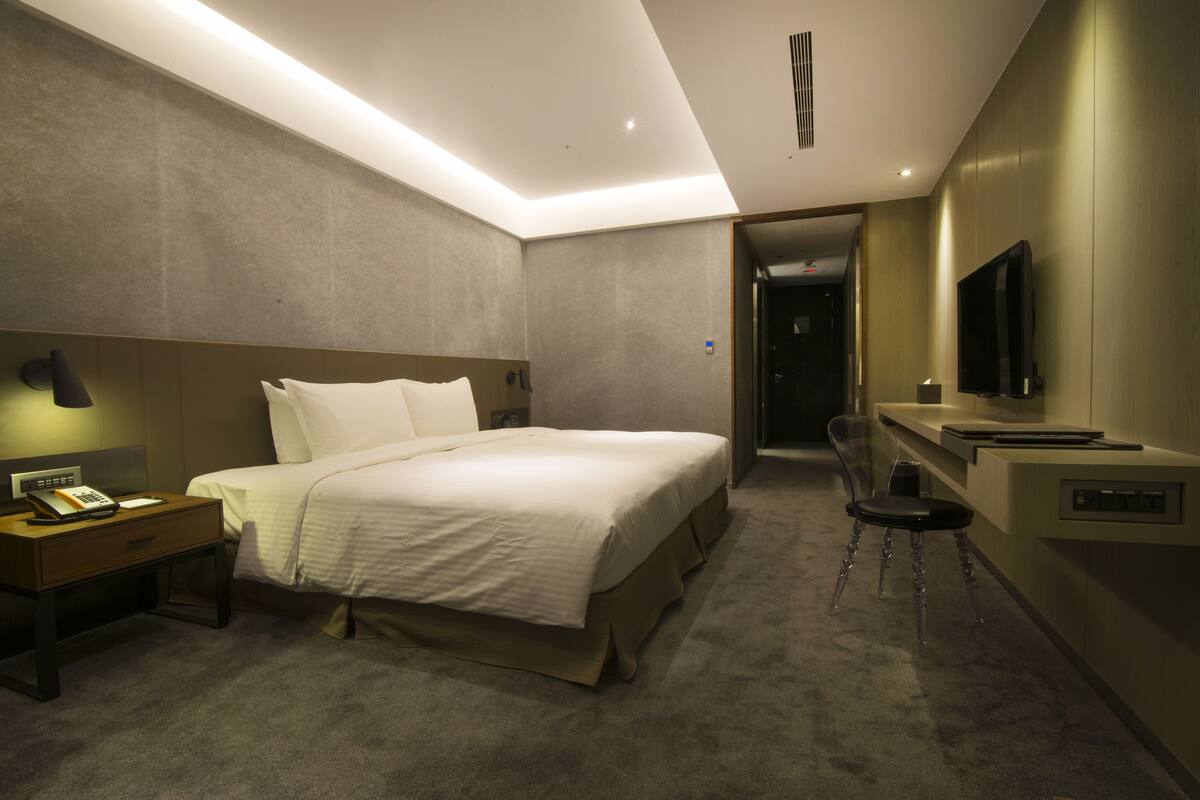 Habitación de hotel con una cama grande, mesita de noche, lámpara, televisión y un escritorio con silla.