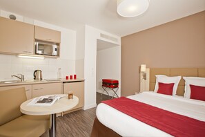 A hotel room with a bed, a small table, a kitchenette, and a bathroom.