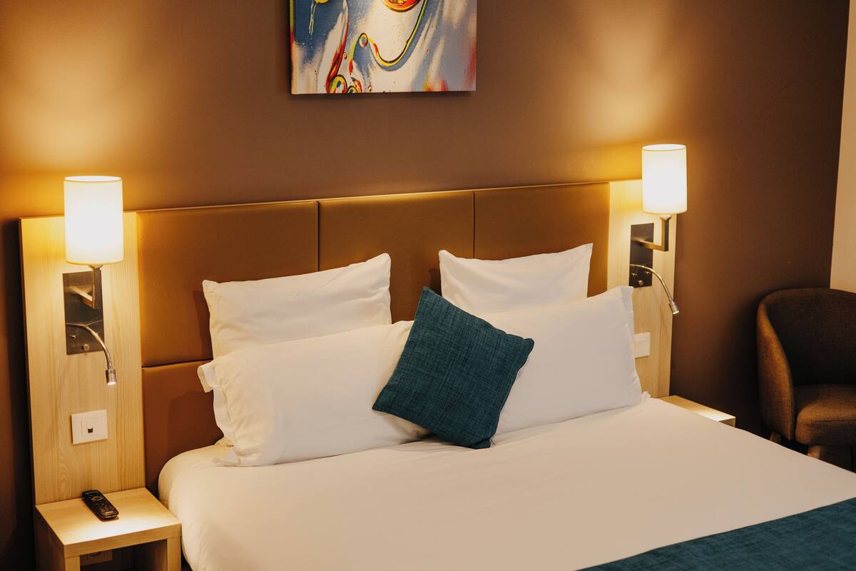 Una habitación de hotel con una cama grande, dos lámparas de noche, un control remoto en la mesita y un cuadro en la pared.