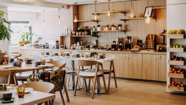 Un café moderno con muebles de madera, lámparas colgantes y una variedad de plantas y frutas en estantes.