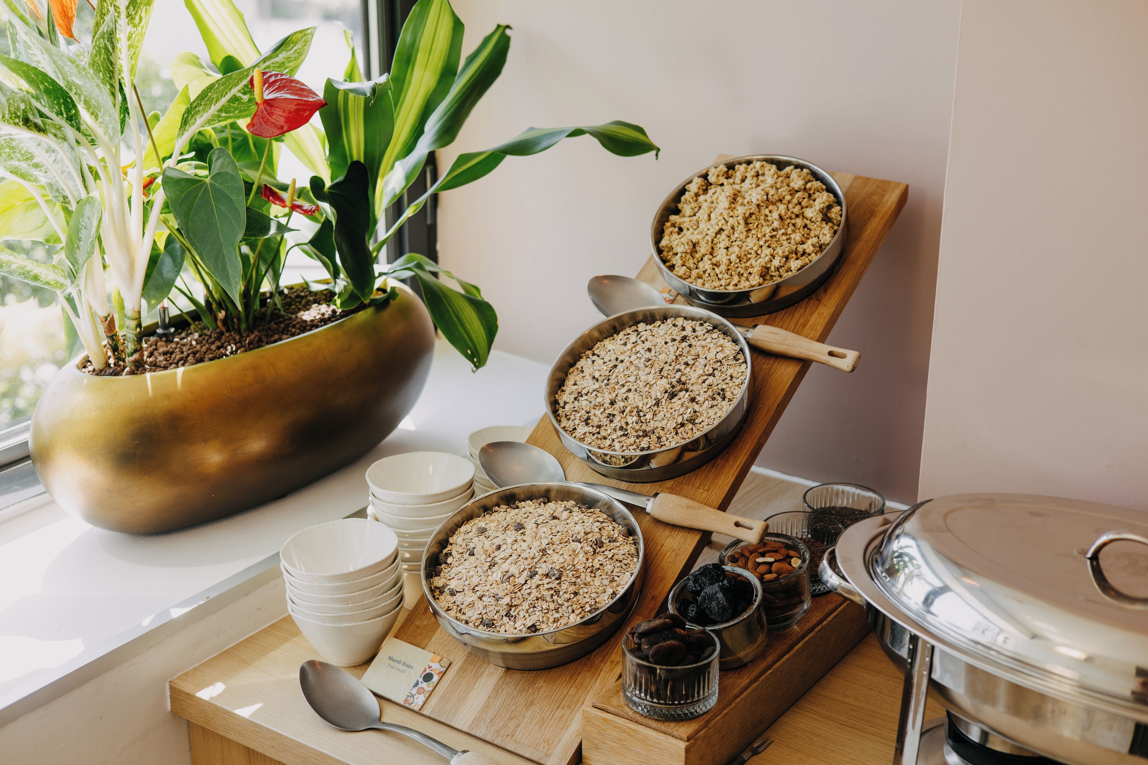 A breakfast buffet with various dishes, including cereal and fruits, arranged on a wooden table near a window with a plant.