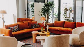 Una sala de estar moderna con sofás anaranjados, un sillón blanco y una mesita de centro. Hay plantas y una lámpara sobre la mesa.