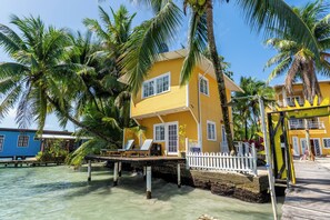 Um edifício amarelo com cerca branca, deck de madeira e palmeiras.