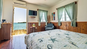 A bedroom with a wooden floor, a bed with a nautical-themed quilt, a ceiling fan, a TV, and a view of the sea through a door. - El Faro del Colibri (Bocas del Toro)