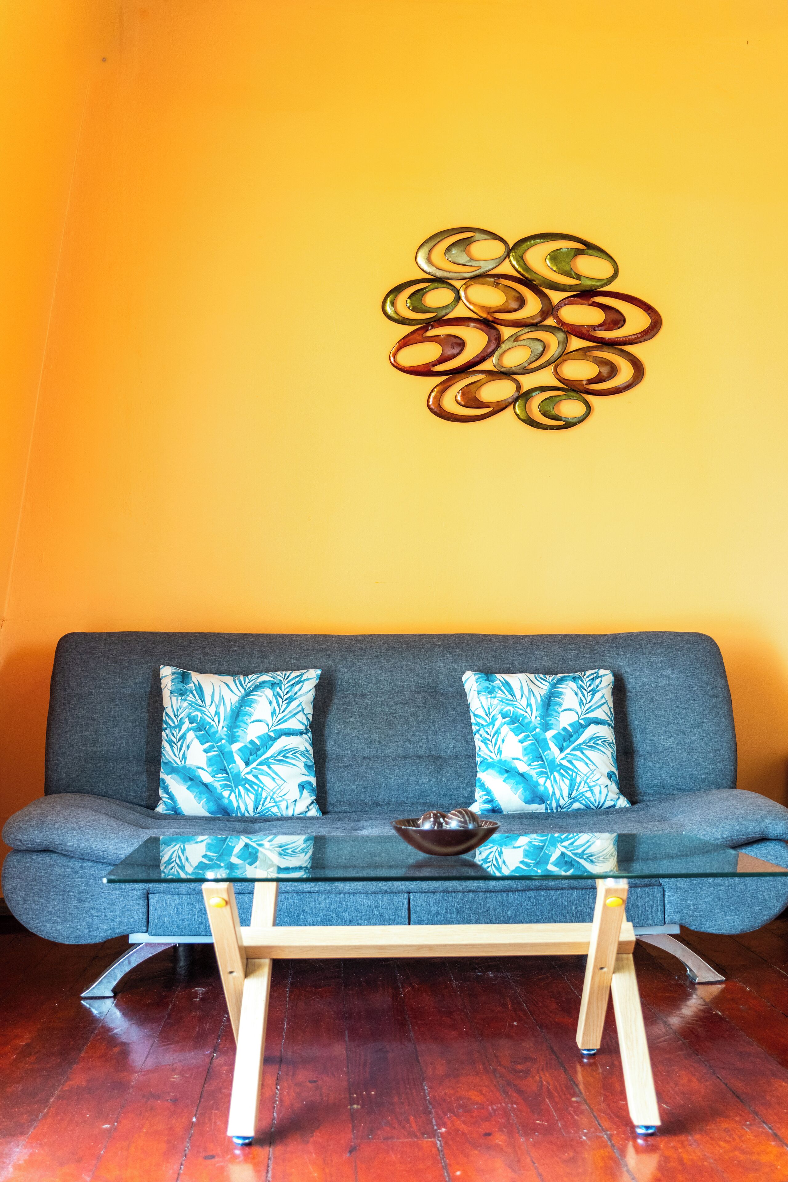 Een kamer met een grijze bank, twee gestreepte kussens, een glazen bijzettafeltje en een decoratief wandobject.