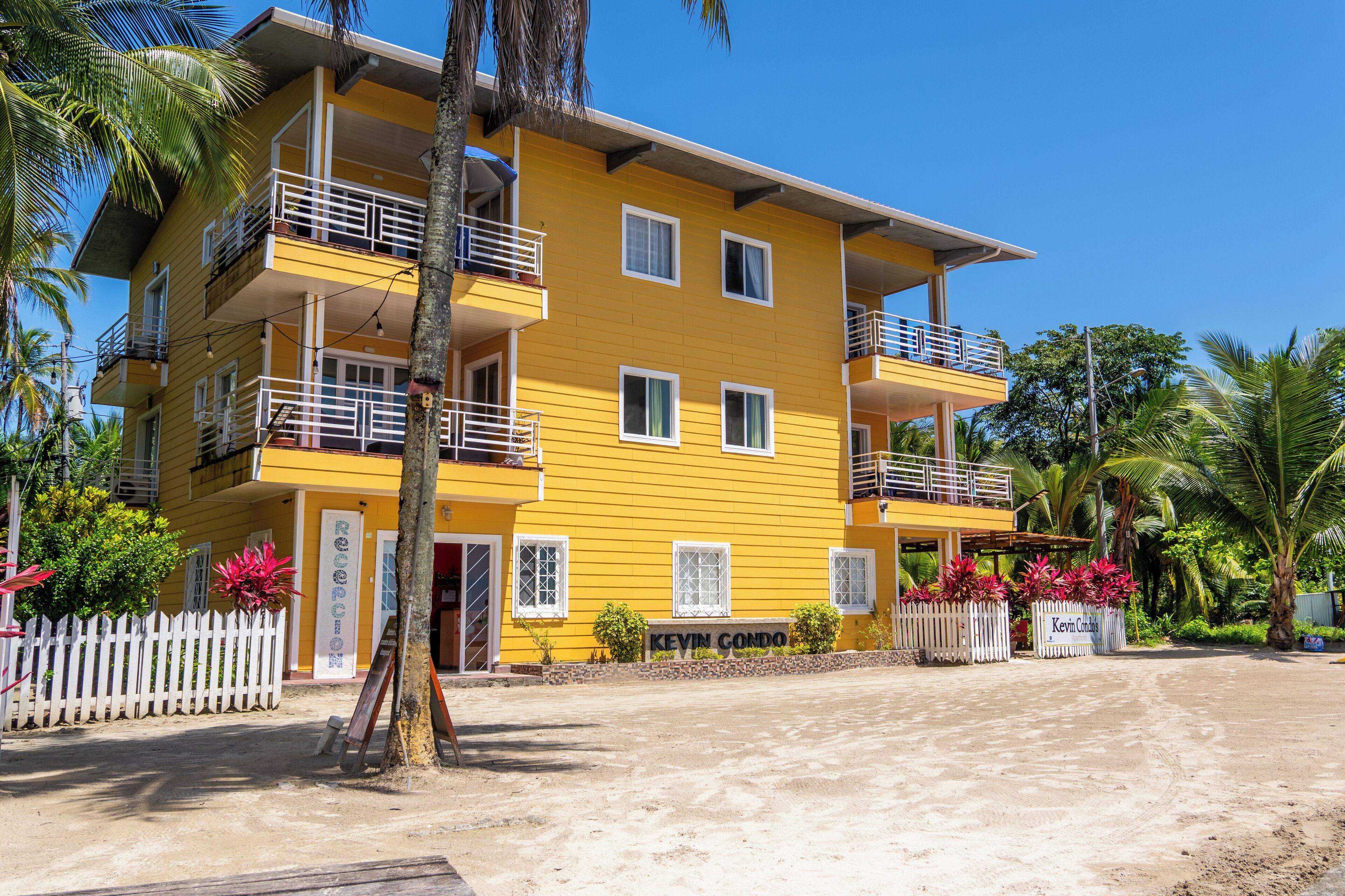 A multi-story yellow building with balconies, a white picket fence, and tropical vegetation.