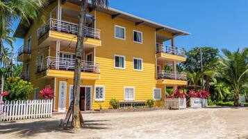 A multi-story yellow building with balconies, a white picket fence, and tropical vegetation.