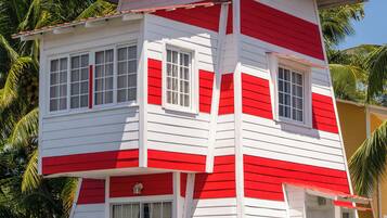 Un bâtiment à plusieurs niveaux, aux murs extérieurs rouges et blancs, construit sur pilotis au-dessus de l’eau et entouré de palmiers.