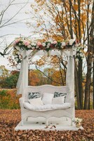 Una silla mecedora de mimbre blanca bajo un arco floral, rodeada de hojas otoñales.