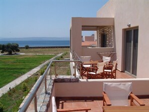 Un balcón con vista al mar, mobiliario exterior y un edificio con techo de tejas.