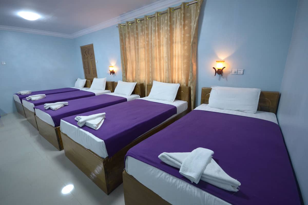 A hotel room with four single beds, each with a purple bedspread and white pillows. There are wall-mounted light fixtures on either side of the window, and a wooden door in the background.