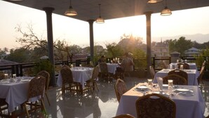 An outdoor dining area with tables set for two, covered by a roof with hanging lights, overlooking a scenic landscape with mountains and trees. - Inlay Palace Hotel (Nyaungshwe)
