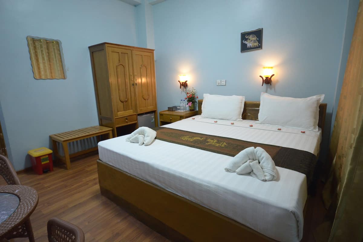 A hotel room with a large bed, wooden wardrobe, bedside tables, a bench, and a small table with chairs.