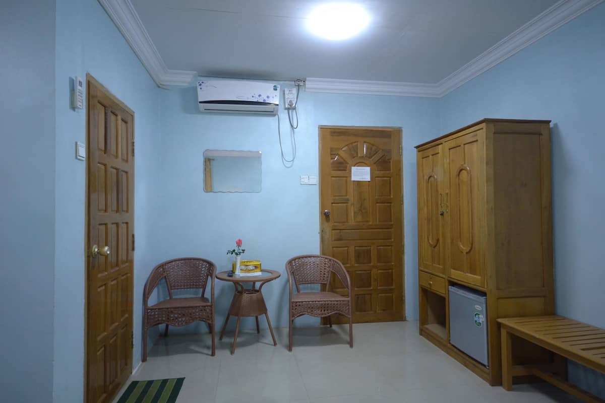A room with two wicker chairs, a small table with a box and a flower, a wooden wardrobe, a bench, and a wall-mounted air conditioner.