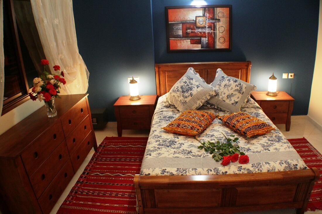 Ein Schlafzimmer mit einem Holzbett, zwei Nachttischen mit Lampen, einer Kommode, einem roten Teppich mit Muster und einem Wandbild.