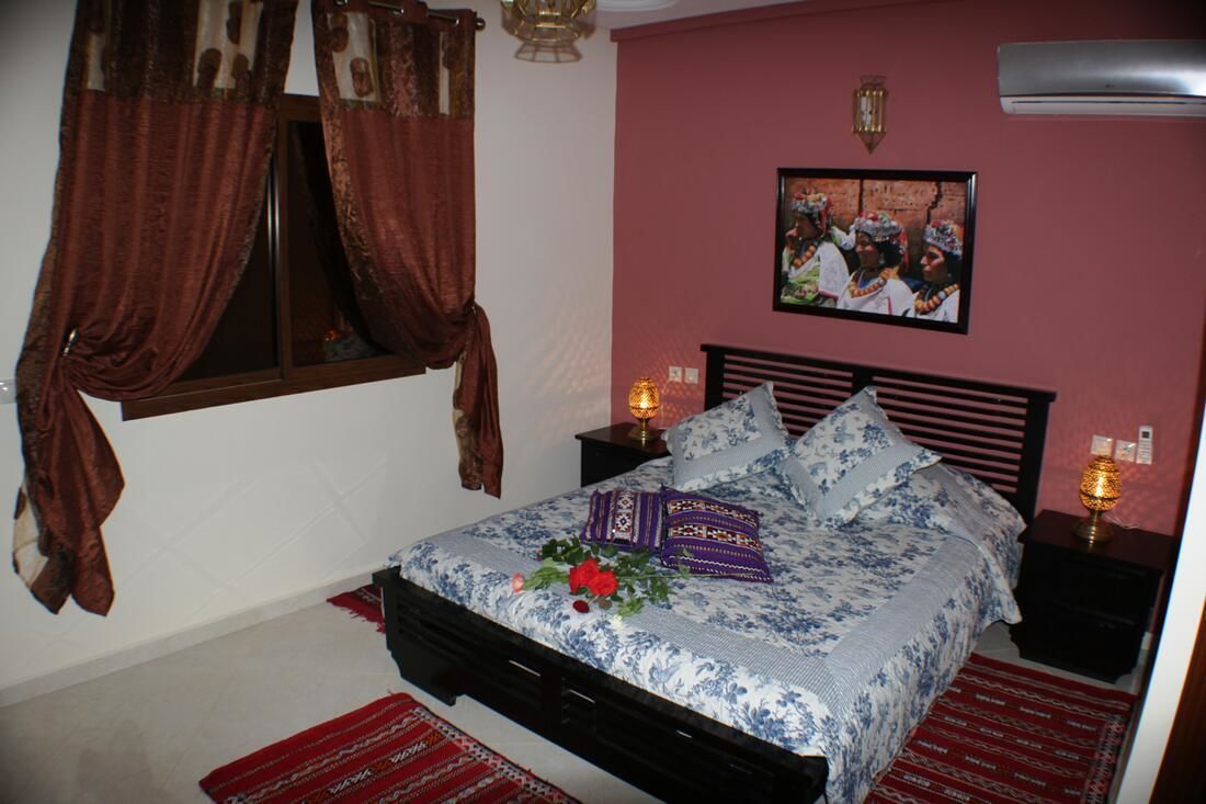 A bedroom with a bed, curtains, a wall-mounted picture, bedside lamps, and a red patterned rug.