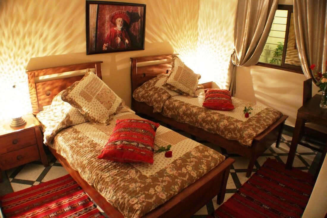 Quarto com duas camas de madeira, cada uma com roupa de cama estampada e almofadas, uma mesa de cabeceira de madeira, um quadro emoldurado na parede e uma janela com cortinas.