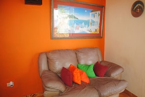 A brown leather sofa with colorful cushions against an orange wall, a framed painting above, and a small decorative plate on the wall. - La Source Hotel & Resort (Roche-à-Bateau)