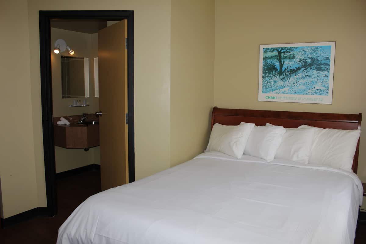 A hotel room with a bed, a wooden headboard, a white wall, a framed picture, and a bathroom visible through an open door.