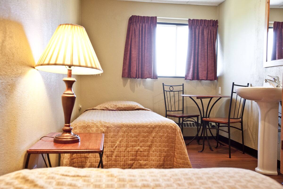 A hotel room with a bed, a desk with a lamp, a chair, a window with curtains, and a sink.