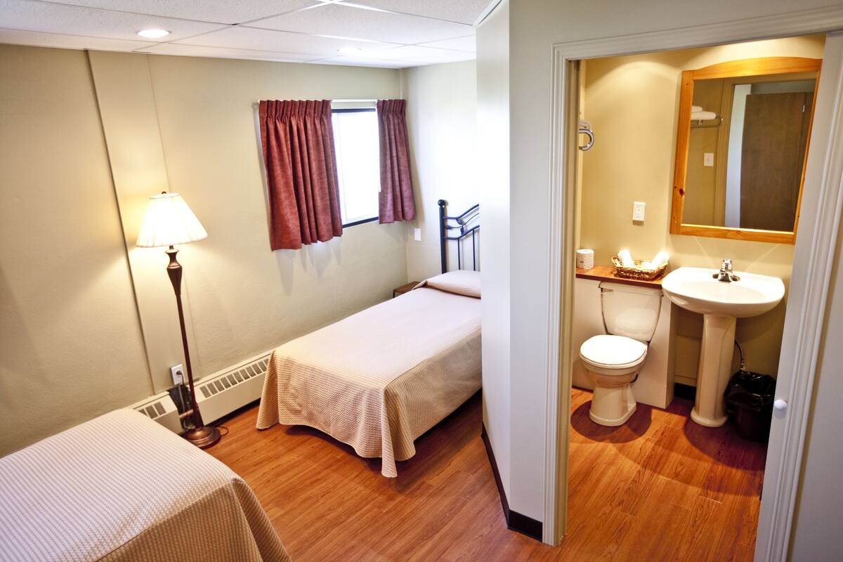 A small, clean room with a single bed, a bedside lamp, a window with curtains, and a bathroom with a sink and toilet.