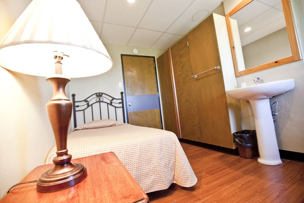 A bedroom with a bed, a bedside table with a lamp, a wooden door, a white sink, and a trash bin.