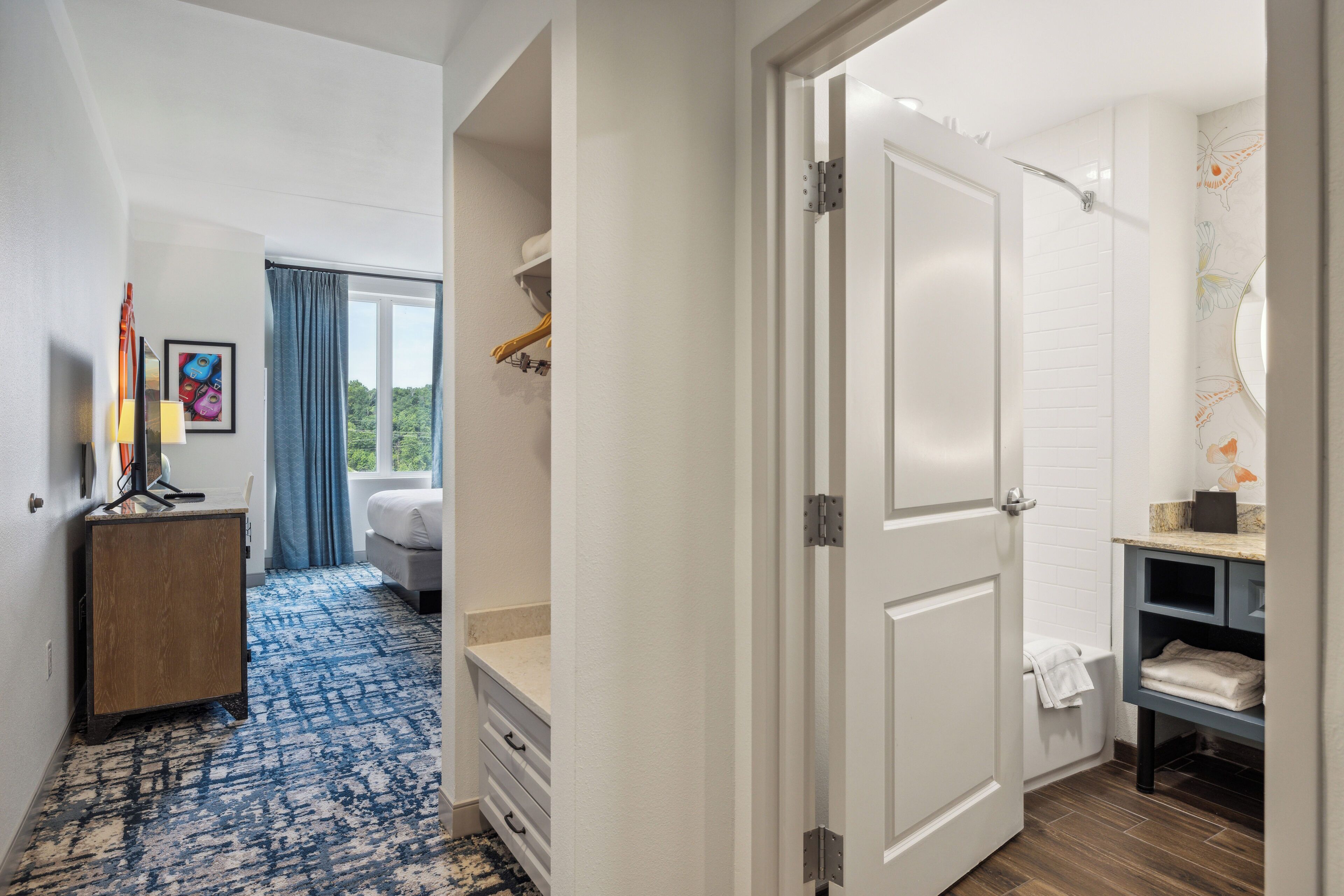 A hotel room with a bed, a desk, a dresser, a bathroom with a shower, and a view of greenery outside.