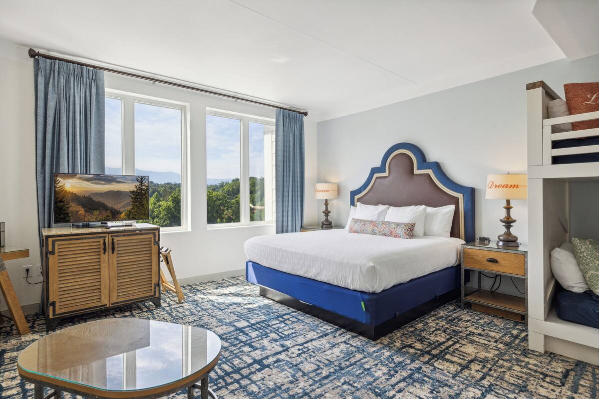 A hotel room with a large bed, a television on a wooden cabinet, a desk with a chair, and a view of the outdoors through the window.